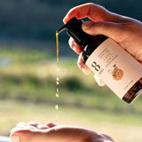 Bottle of '8 Days' body oil with dropper being held over hands against a natural background