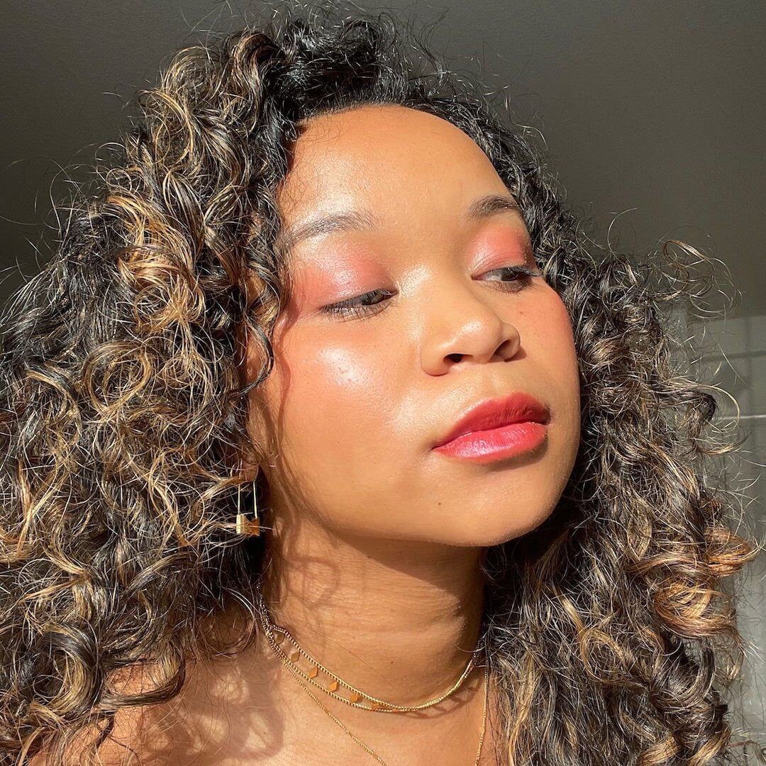 Woman with curly hair and axiology multi-stick in shade joy makeup against a neutral background