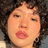 Close-up of a person with curly hair and hoop earrings, wearing Axiology Tinted Dew in shade 'The Goodness'