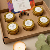 A set of six small soy candles in gold metal tins arranged in a brown cardboard gift box. Each tin has a white label with scent names including Orange + Grape, Rosemary + Lemon, Eucalyptus + Lavender, and Mint + Garden.
