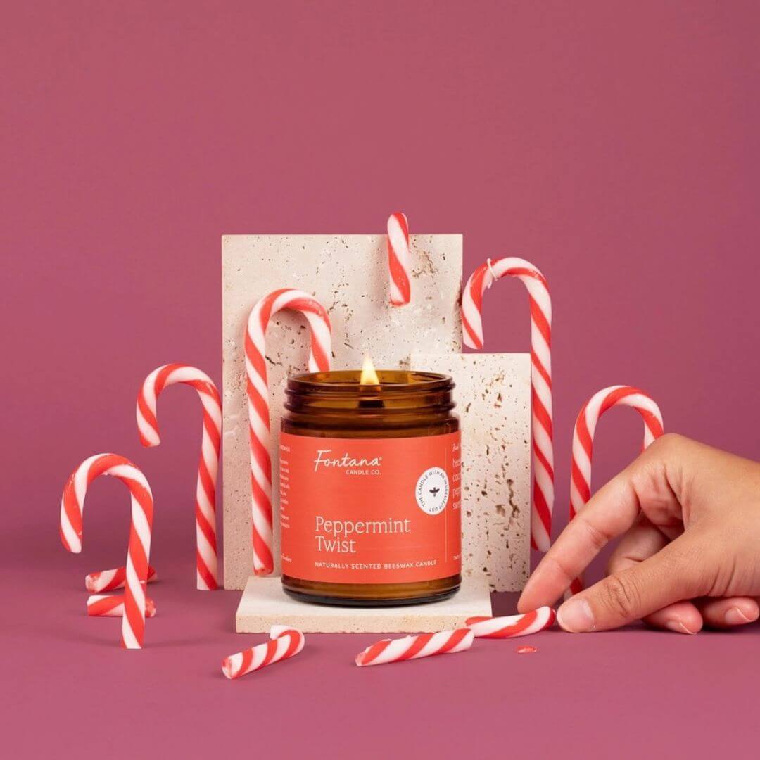 An amber glass jar candle labeled 'Peppermint Twist' with a lit flame, styled with red-and-white striped candy canes on a dusty pink background. A human hand is visible at the right edge, and the candle is set against white textured display blocks.