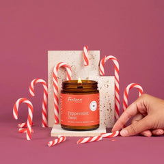 An amber glass jar candle labeled 'Peppermint Twist' with a lit flame, styled with red-and-white striped candy canes on a dusty pink background. A human hand is visible at the right edge, and the candle is set against white textured display blocks.