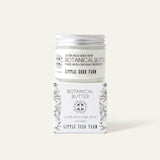 A jar of Little Seed Farm Botanical Butter shea whipped cream is stacked on top of its matching illustrated cardboard box against a solid light beige background.
