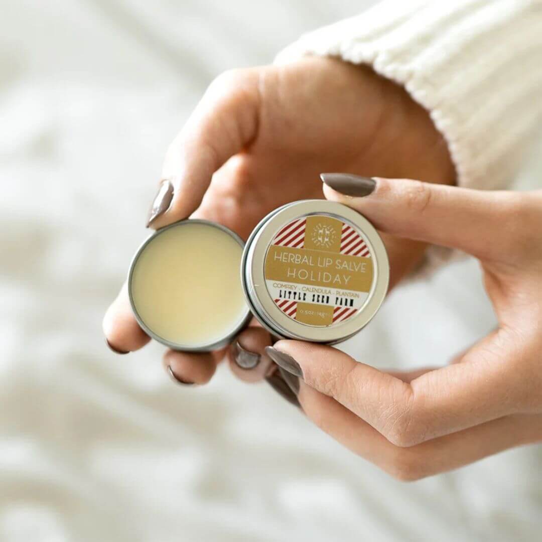A close-up of a person's hands holding an open round tin of herbal lip balm, showing the pale yellow balm inside and the lid label that reads 'HERBAL LIP BALM HOLIDAY'.