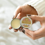 A close-up of a person's hands holding an open round tin of herbal lip balm, showing the pale yellow balm inside and the lid label that reads 'HERBAL LIP BALM HOLIDAY'.