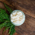 A jar filled with Little Seed Farm Organic  exfoliating scrub, photographed from above on a rustic wooden surface with green pine branches around it.