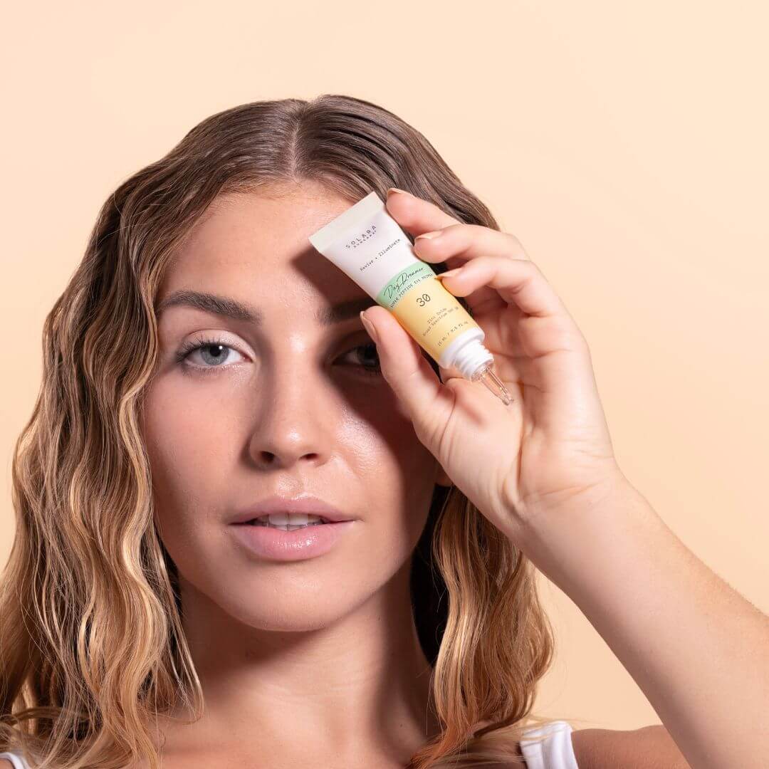 Woman holding tube of solara day dreamer peptide eye cream to her face with a beige background