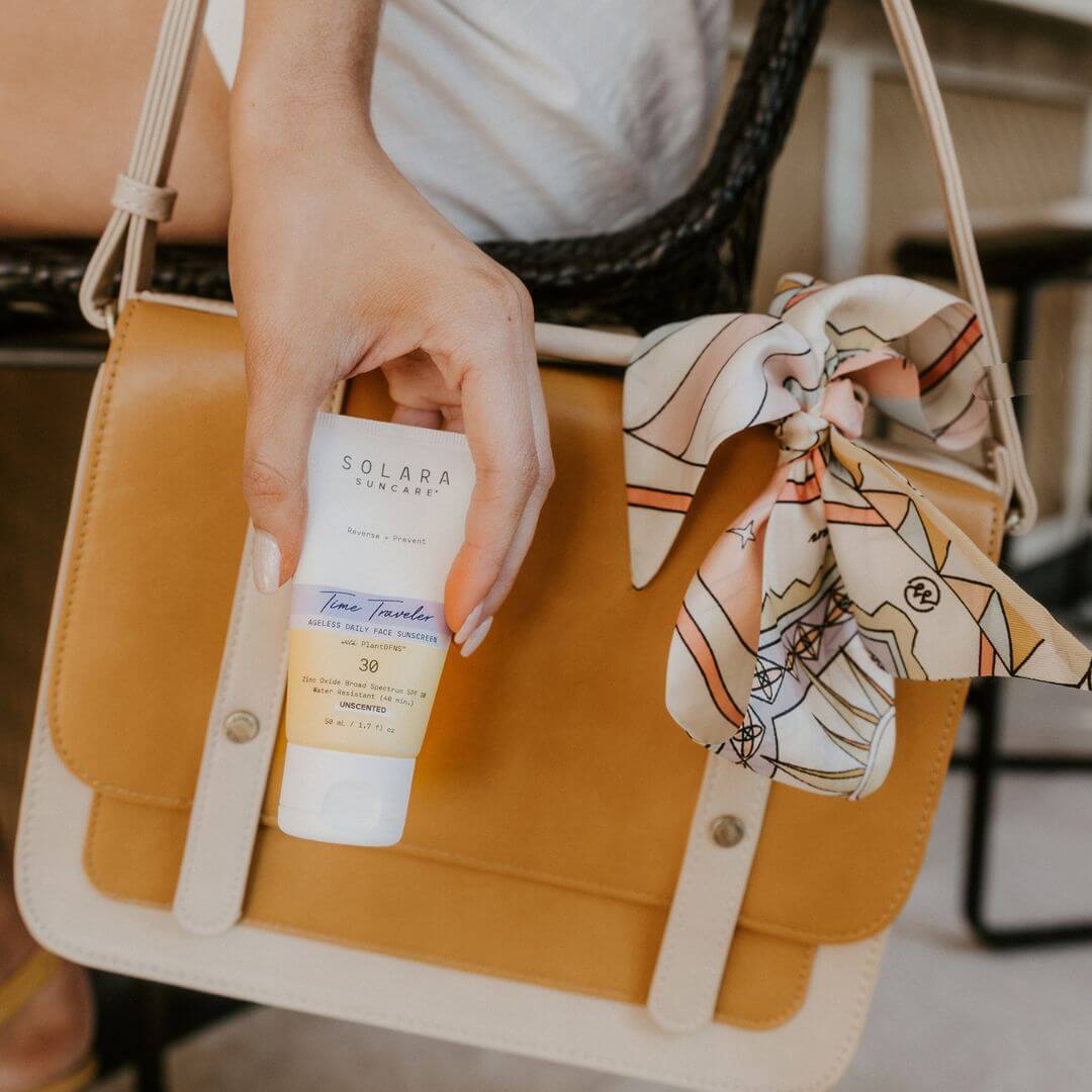 Person holding a bottle of Solara Time Traveler sunscreen next to a yellow handbag with a decorative bow.