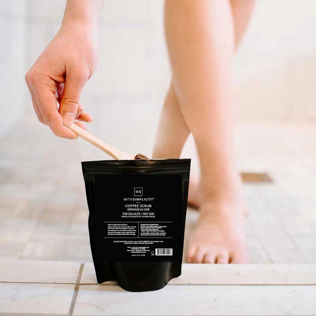 Person using a coffee scrub packet on their leg with a wooden applicator.