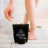 Person using a coffee scrub packet on their leg with a wooden applicator.