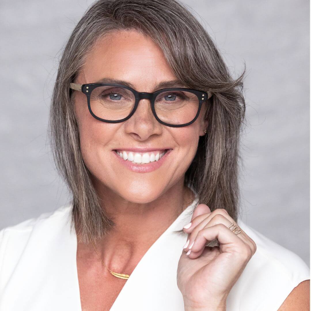 Woman with gray hair wearing glasses and a white shirt against a gray background