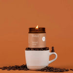A lit candle jar labeled 'Fontana Candle Co. Spiced Latte' sits on top of a white ceramic mug, surrounded by scattered coffee beans against a solid warm orange-brown background.