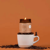 A lit candle jar labeled 'Fontana Candle Co. Spiced Latte' sits on top of a white ceramic mug, surrounded by scattered coffee beans against a solid warm orange-brown background.