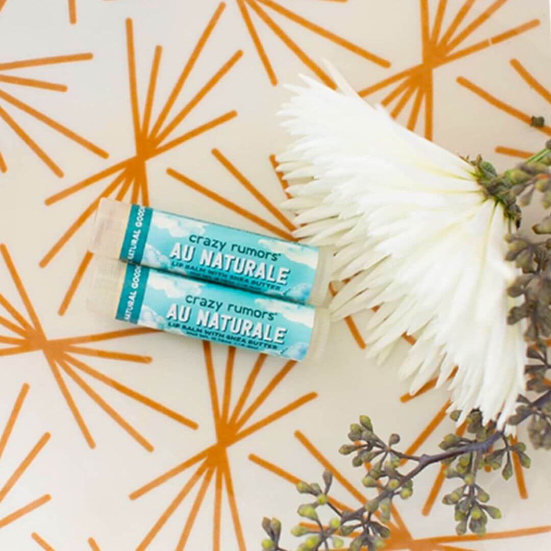 Two tubes of 'crazy rumors Au Naturale' lip balm on a decorative background with orange abstract patterns and green leaves.