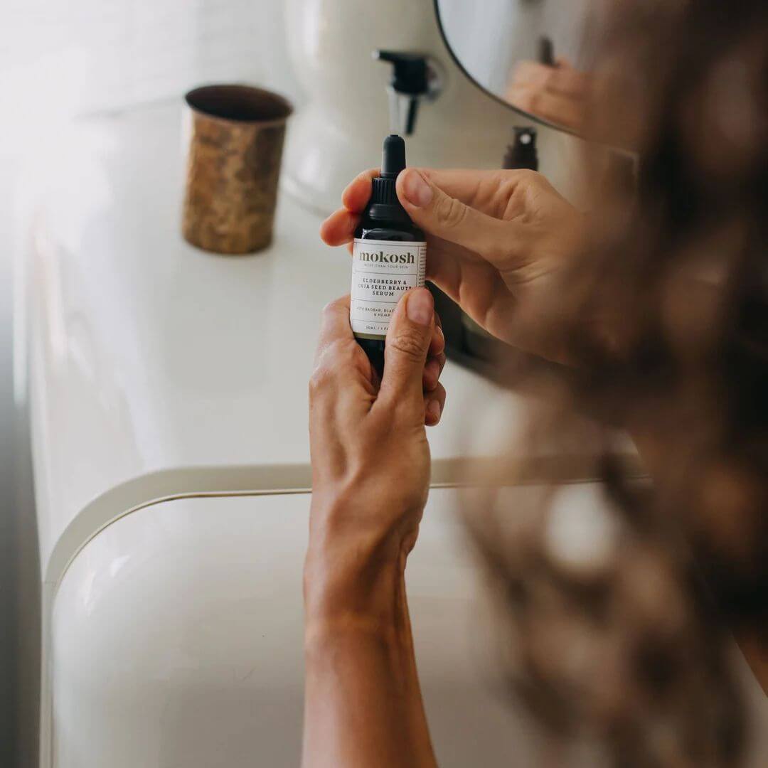 Person holding a bottle of mokosh skincare serum in front of a mirror.