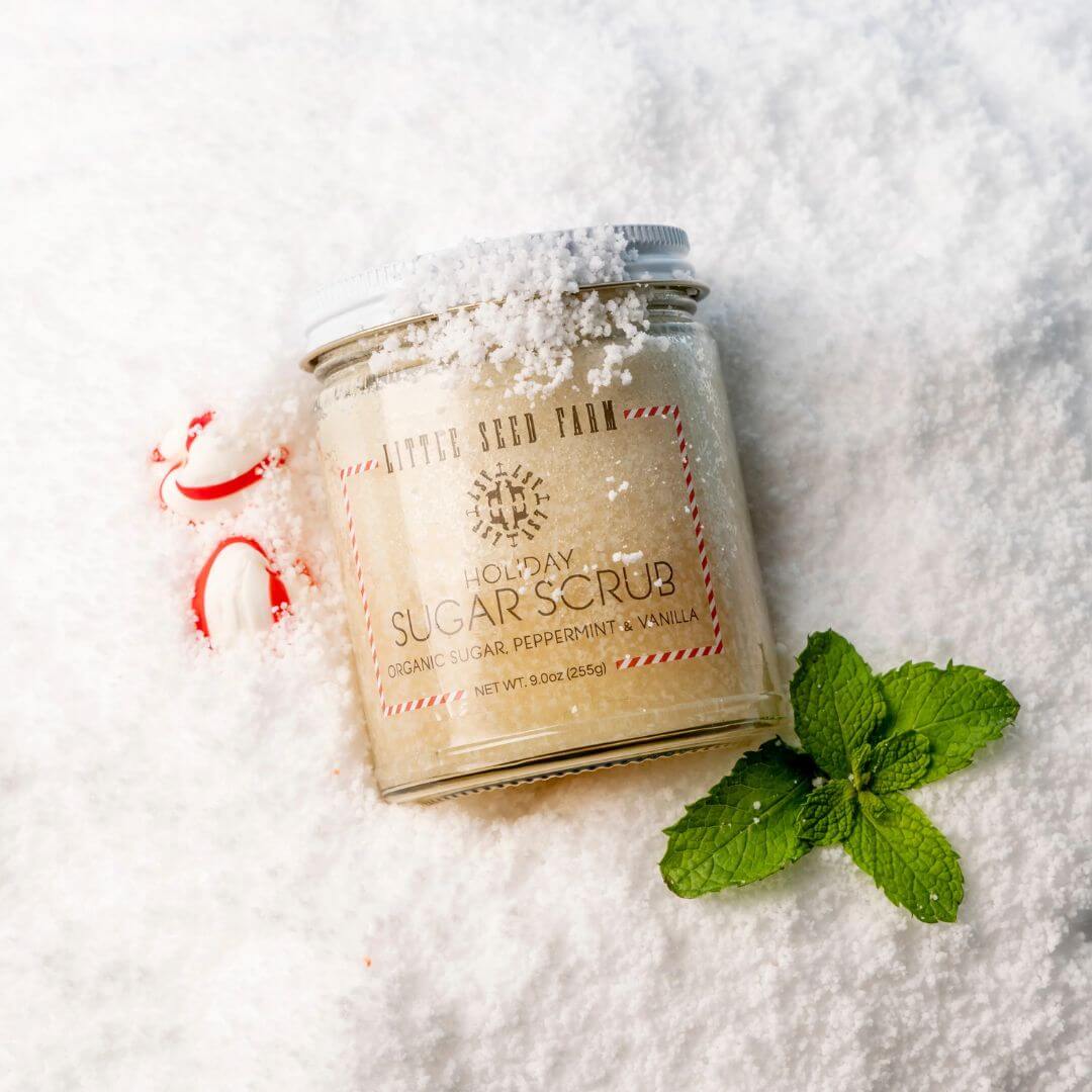 A jar of Little Seed Farm Holiday Sugar Scrub, scented with peppermint and vanilla, styled on a bed of white sugar with mint leaves and candy canes.