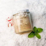 A jar of Little Seed Farm Holiday Sugar Scrub, scented with peppermint and vanilla, styled on a bed of white sugar with mint leaves and candy canes.