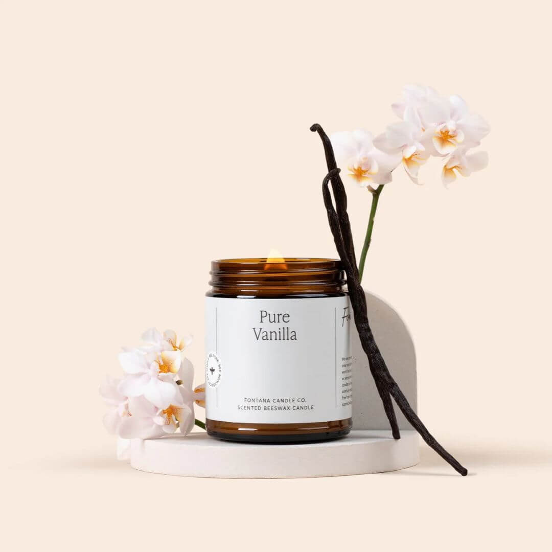 An amber glass jar candle labeled 'Pure Vanilla' by Fontana Candle Co., styled on a white pedestal with white flowers and vanilla beans against a warm beige background.