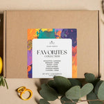 A kraft paper gift box with a colorful watercolor-style label reading 'SLOW NORTH FAVORITES COLLECTION' and listing candle scents, styled on a beige surface with green leaves and small yellow rings.