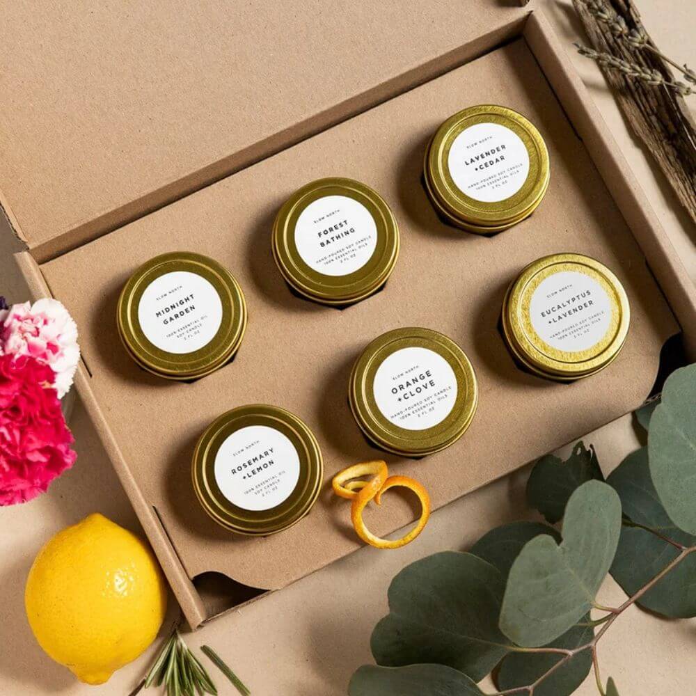 A gift set of six small beeswax candles in gold metal tins arranged inside a brown kraft cardboard box. The candles have white labels with black text, including scents like Honey Balm, Rosemary Lemon, and Eucalyptus Aromat.