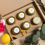 A gift set of six small beeswax candles in gold metal tins arranged inside a brown kraft cardboard box. The candles have white labels with black text, including scents like Honey Balm, Rosemary Lemon, and Eucalyptus Aromat.
