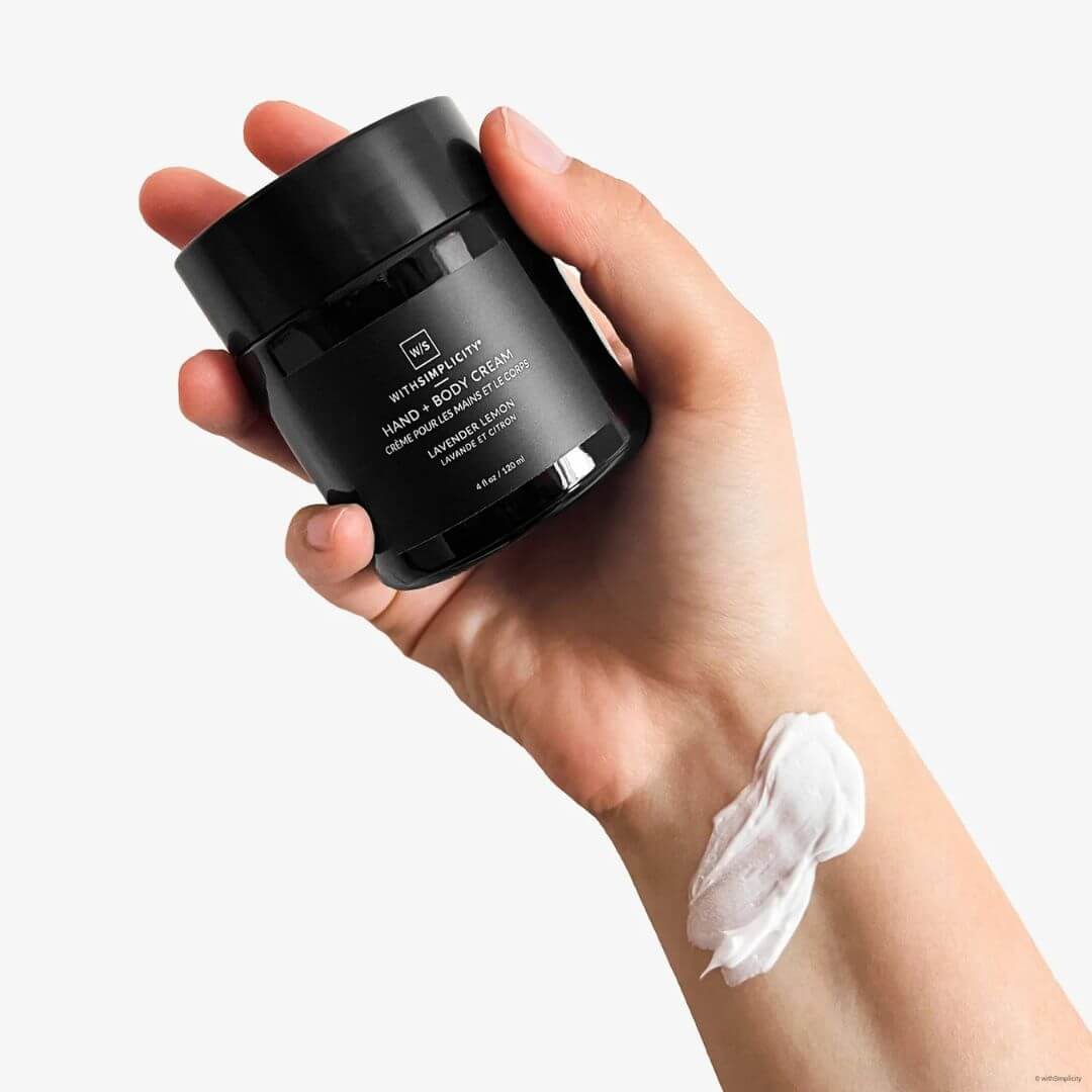Hand holding a black jar of withSimplcity body cream, showing cream on a wrist against a white background.
